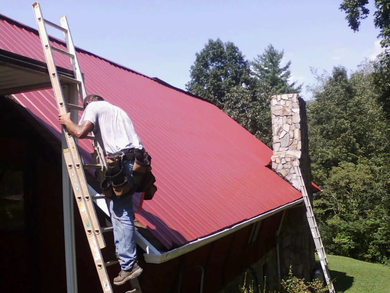 Roofer on a ladder beside a red metal roof and stone chimney — Soffit & Fascia Repair by Roofing Brothers in Riddleton